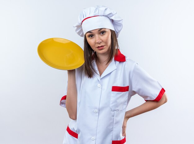 Confiado joven cocinera vistiendo uniforme de chef sosteniendo la placa poniendo la mano en la cadera aislado sobre fondo blanco.