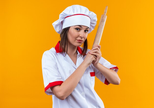 Confiado joven cocinera vistiendo uniforme de chef sosteniendo un rodillo aislado sobre fondo naranja