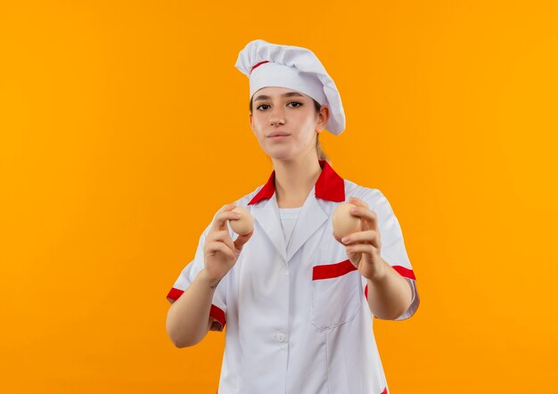 Confiado joven cocinero bonito en uniforme de chef sosteniendo y estirando huevos aislados en la pared naranja con espacio de copia