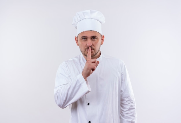 Foto gratuita confiado joven cocinero guapo en uniforme de chef gesticulando silencio aislado en la pared blanca con espacio de copia