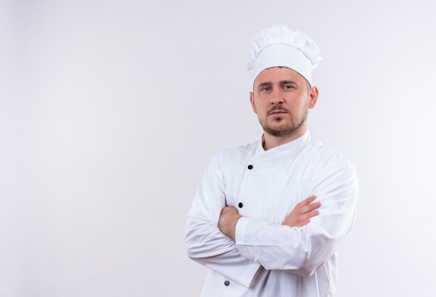Confiado joven cocinero guapo en uniforme de chef de pie con postura cerrada aislada en la pared blanca