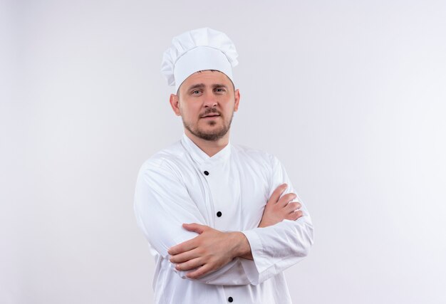 Confiado joven cocinero guapo en uniforme de chef de pie con postura cerrada aislada en la pared blanca