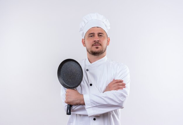 Confiado joven cocinero guapo en uniforme de chef de pie con postura cerrada sosteniendo una sartén aislada en la pared blanca