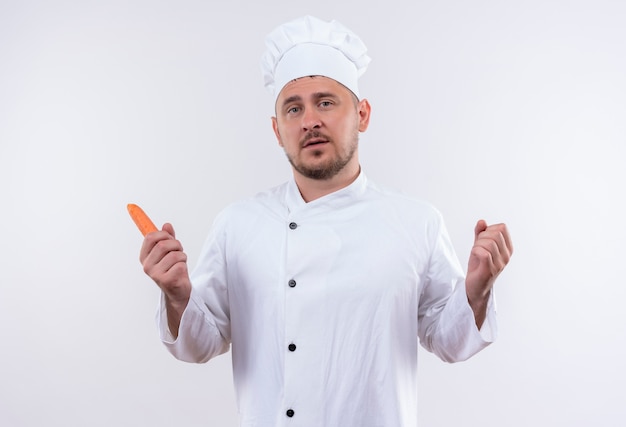 Confiado joven cocinero guapo en uniforme de chef con zanahoria aislado en la pared blanca