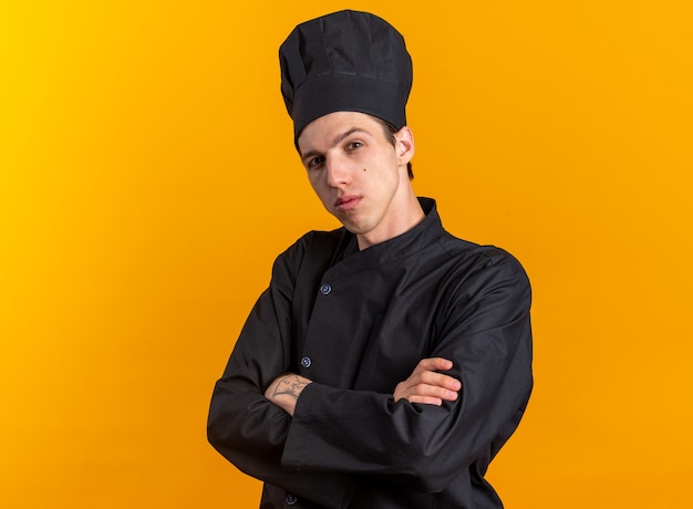 Confiado joven cocinero rubio en uniforme de chef y gorra de pie en vista de perfil con postura cerrada mirando a cámara aislada en pared naranja con espacio de copia