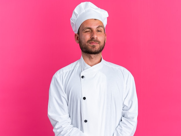 Confiado joven cocinero de sexo masculino caucásico en uniforme de chef y gorra manteniendo las manos juntas mirando a la cámara guiñando un ojo aislado en la pared rosa