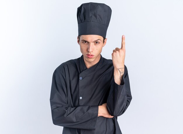 Confiado joven cocinero de sexo masculino rubio en uniforme de chef y gorra mirando a cámara mirando a cámara apuntando hacia arriba aislado en la pared blanca con espacio de copia