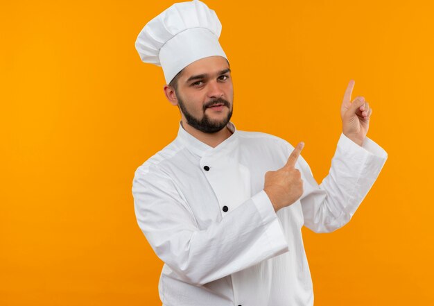 Confiado joven cocinero en uniforme de chef apuntando hacia arriba aislado en la pared naranja con espacio de copia