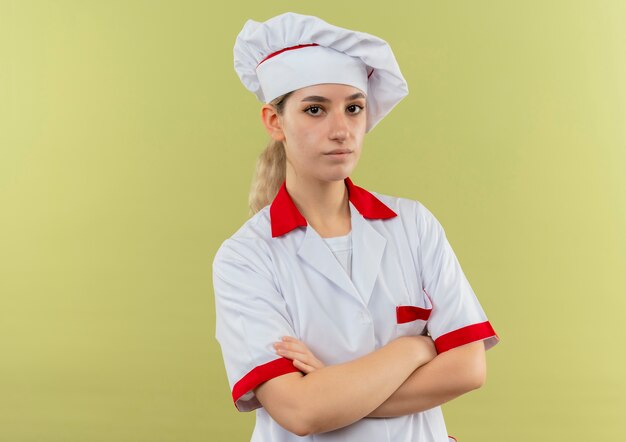 Confiado joven cocinero en uniforme de chef de pie con postura cerrada aislada en la pared verde con espacio de copia