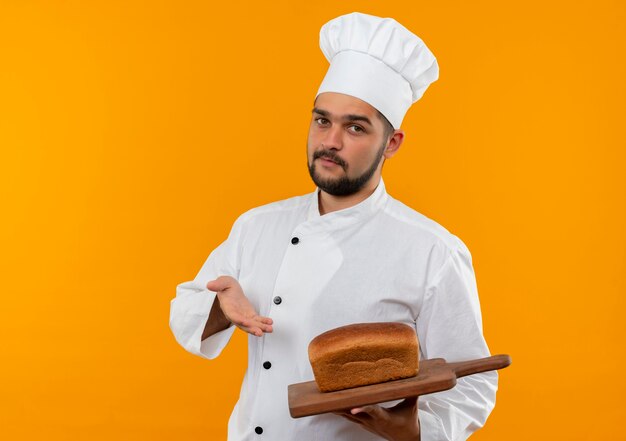 Confiado joven cocinero en uniforme de chef sosteniendo y apuntando con la mano a la tabla de cortar con pan aislado en la pared naranja