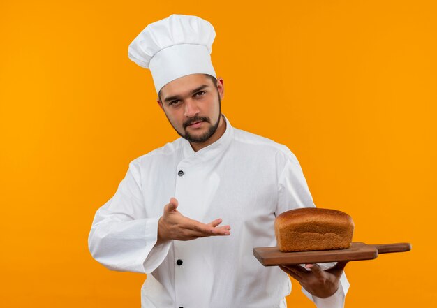Confiado joven cocinero en uniforme de chef sosteniendo y apuntando con la mano a la tabla de cortar con pan aislado en la pared naranja