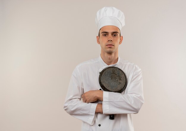 Confiado joven cocinero vistiendo uniforme de chef cruzando las manos y sosteniendo una sartén en la pared blanca aislada con espacio de copia