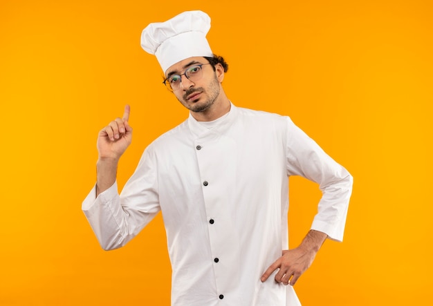 Confiado joven cocinero vistiendo uniforme de chef y gafas apunta hacia arriba y poniendo la mano en la cadera