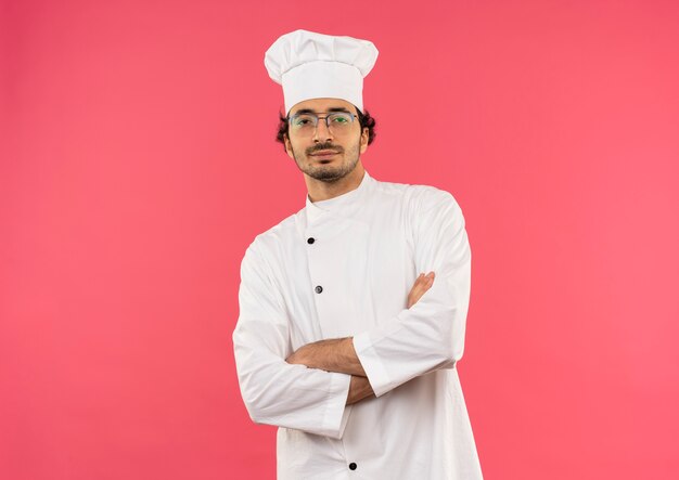 Confiado joven cocinero vistiendo uniforme de chef y gafas cruzando las manos