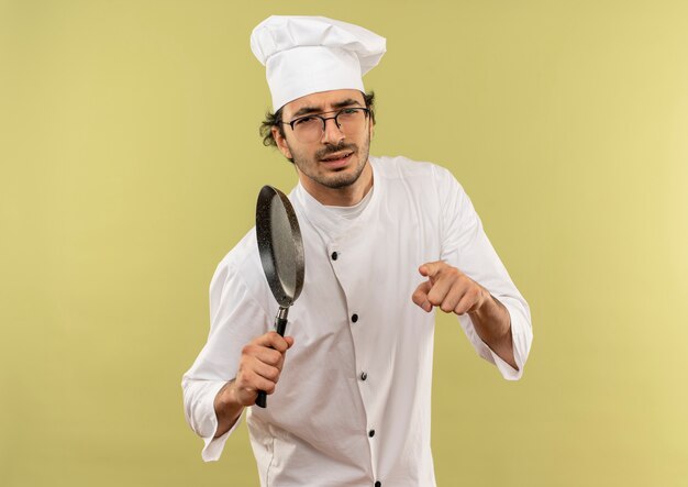 Confiado joven cocinero vistiendo uniforme de chef y gafas sosteniendo una sartén y mostrando su gesto