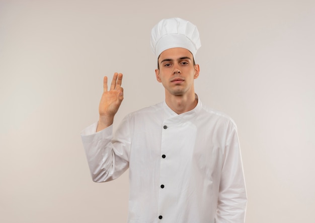 Confiado joven cocinero vistiendo uniforme de chef mostrando gesto okey en pared blanca aislada con espacio de copia