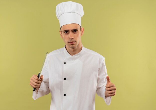 Confiado joven cocinero vistiendo uniforme de chef sosteniendo la cuchilla con el pulgar hacia arriba en la pared verde aislada con espacio de copia