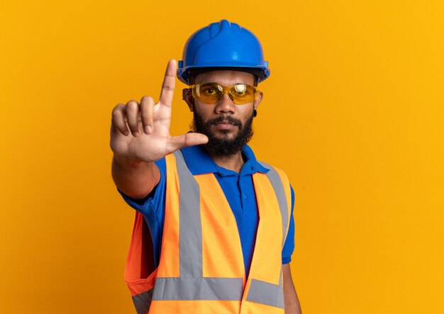 Confiado joven constructor afroamericano en gafas de seguridad con uniforme con casco de seguridad apuntando hacia arriba aislado en la pared naranja con espacio de copia