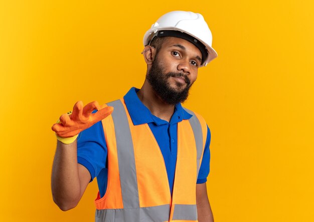 Confiado joven constructor afroamericano en uniforme con casco de seguridad y guantes manteniendo su mano abierta aislada sobre fondo naranja con espacio de copia