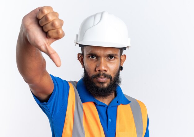 Confiado joven constructor afroamericano en uniforme con casco de seguridad pulgar hacia abajo aislado sobre fondo blanco con espacio de copia