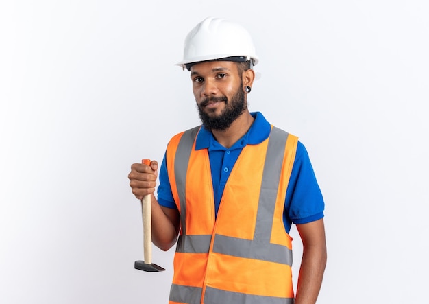 Confiado joven constructor afroamericano en uniforme con casco de seguridad sosteniendo el martillo boca abajo aislado en la pared blanca con espacio de copia