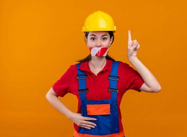 Confiado joven constructor boca sellada con cinta de advertencia y apunta hacia arriba sobre fondo naranja aislado