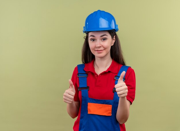 Confiado joven constructor con casco de seguridad azul pulgar hacia arriba con ambas manos sobre fondo verde aislado con espacio de copia