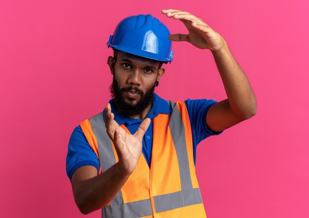 Confiado joven constructor hombre en uniforme con casco de seguridad fingiendo sostener algo aislado en la pared rosa con espacio de copia