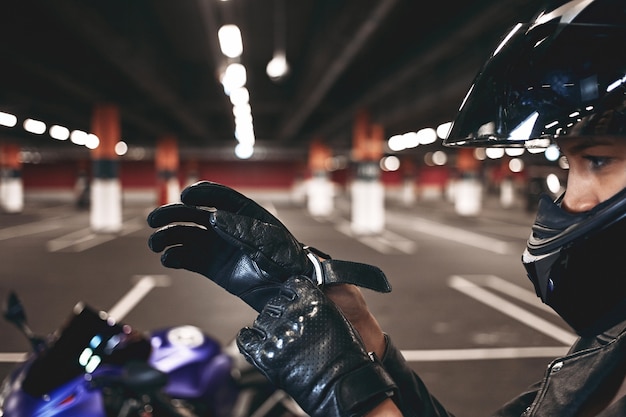 Confiado joven corredor con elegante casco de motocicleta poniéndose guantes de cuero, posando aislado en el estacionamiento subterráneo con su moto azul. Enfoque selectivo en manos de mujer