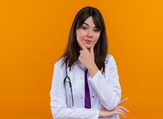 Confiado joven doctora en bata médica con estetoscopio pone la mano en la barbilla sobre fondo naranja aislado con espacio de copia
