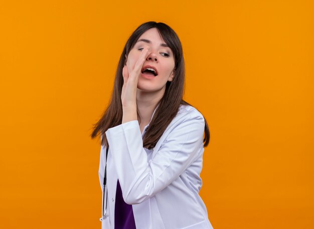 Confiado joven doctora en bata médica con estetoscopio pretende llamar a alguien sobre fondo naranja aislado con espacio de copia