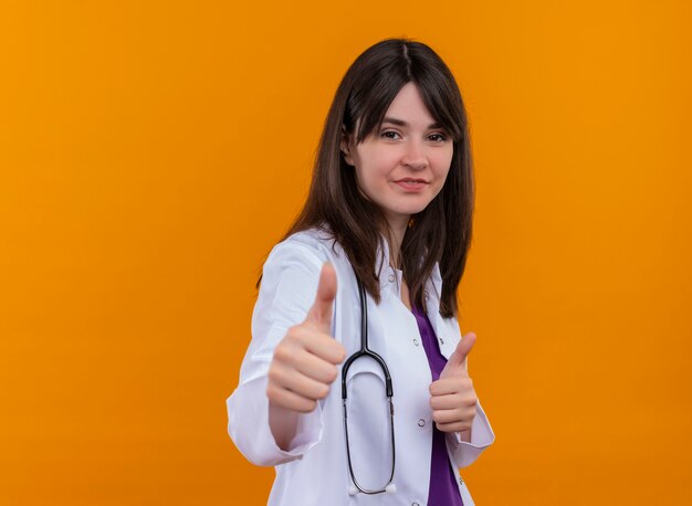 Confiado joven doctora en bata médica con estetoscopio pulgar hacia arriba sobre fondo naranja aislado con espacio de copia