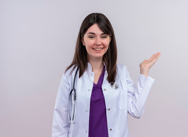 Confiado joven doctora en bata médica con estetoscopio sostiene la mano vacía sobre fondo blanco aislado con espacio de copia