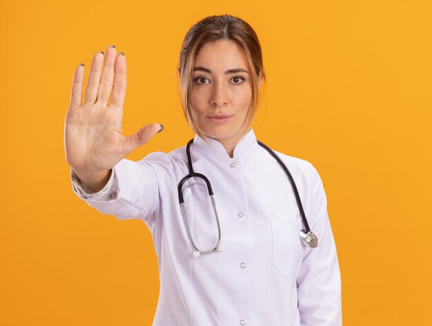 Confiado joven doctora vistiendo bata médica con estetoscopio mostrando gesto de parada aislado en la pared amarilla