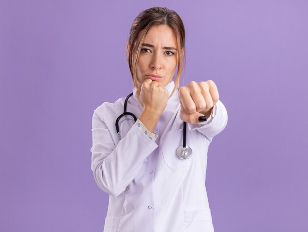 Confiado joven doctora vistiendo bata médica con estetoscopio sosteniendo el puño a la cámara aislada en la pared púrpura
