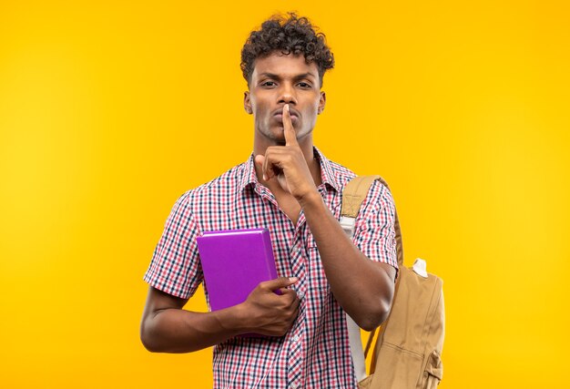 Confiado joven estudiante afroamericano con mochila sosteniendo el libro y haciendo gesto de silencio aislado en la pared naranja con espacio de copia