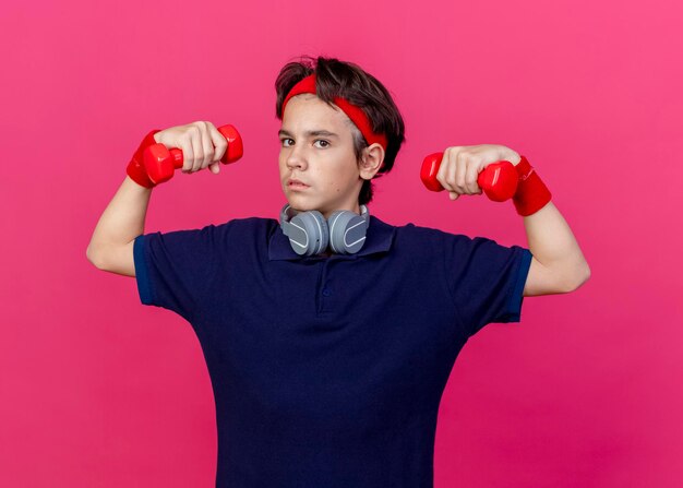 Confiado joven guapo deportivo con diadema y muñequeras y auriculares en el cuello con aparatos dentales levantando pesas mirando al frente aislado en la pared rosa