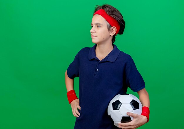 Confiado joven guapo deportivo vistiendo diadema y muñequeras con aparatos dentales sosteniendo un balón de fútbol manteniendo la mano en la cintura mirando directamente aislado en la pared verde con espacio de copia