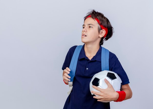 Foto gratuita confiado joven guapo deportivo vistiendo diadema y muñequeras y bolso trasero con aparatos dentales que sostienen la correa del bolso y el balón de fútbol mirando al lado aislado sobre fondo blanco con espacio de copia