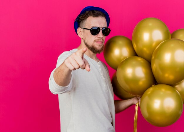 Confiado joven guapo partido eslavo con sombrero de fiesta y gafas de sol sosteniendo globos mirando y apuntando al frente aislado en la pared rosa con espacio de copia