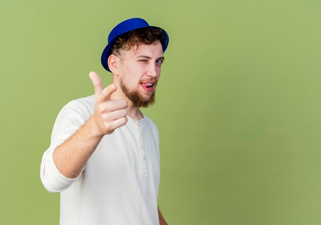 Confiado joven guapo partido eslavo con sombrero de fiesta guiñando un ojo mostrando la lengua mirando y apuntando a la cámara aislada sobre fondo verde oliva con espacio de copia