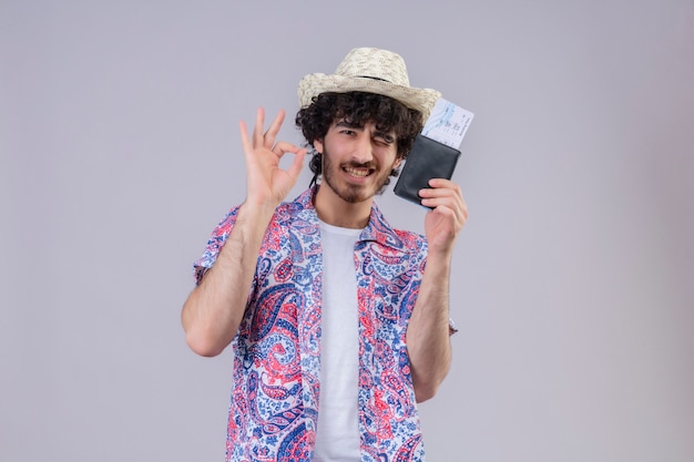 Confiado joven guapo viajero rizado hombre con sombrero haciendo el signo de ok, mostrando billetera y boletos de avión en un espacio en blanco aislado con espacio de copia