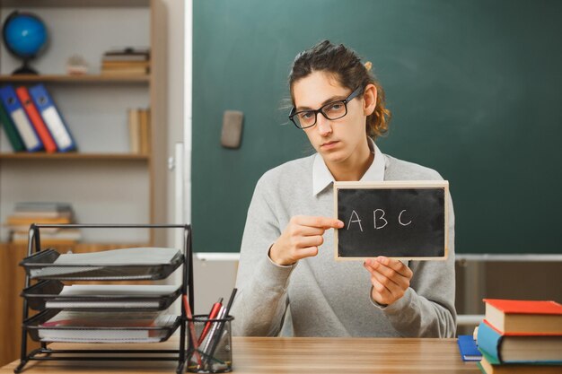 confiado joven maestro usando anteojos sosteniendo y apuntando a la mini pizarra sentado en el escritorio con herramientas escolares en el aula