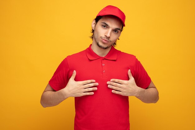 confiado joven repartidor con uniforme y gorra mirando a la cámara apuntándose a sí mismo con las manos aisladas en el fondo amarillo