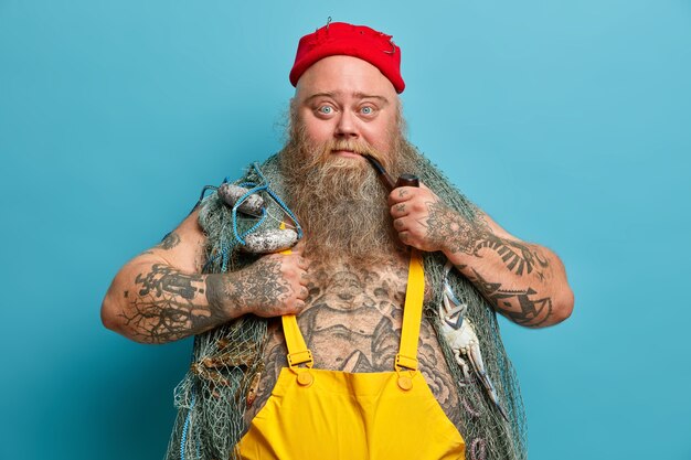 Confiado marinero barbudo mira seriamente a la cámara fuma pipa de tabaco y posa con red de pesca tiene tatuajes usa sombrero rojo con anzuelos disfruta de su pasatiempo favorito
