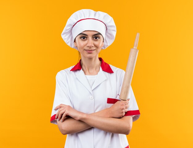 Confiado mirando a la cámara hermosa joven en uniforme de chef sosteniendo el rodillo cruzando las manos