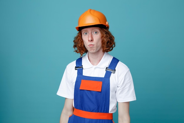 Confiado mirando a la cámara joven constructor hombre en uniforme aislado sobre fondo azul.