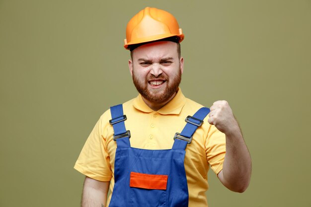 Confiado mostrando gesto fuerte joven constructor hombre en uniforme aislado sobre fondo verde