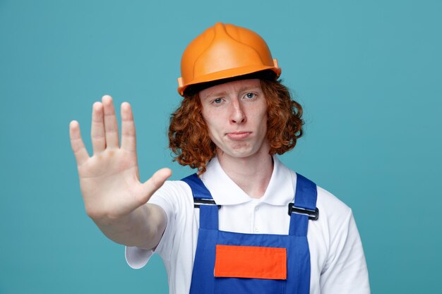 Confiado mostrando gesto de parada joven constructor hombre en uniforme aislado sobre fondo azul.