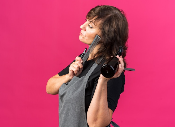 Foto gratuito confiado peluquería femenina adulta en uniforme de pie lateralmente sosteniendo herramientas de peluquería aislado en la pared rosa con espacio de copia